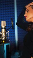 magnifique femme avec longue cheveux est en chantant et dansant près le microphone dans le l'audio studio sur une bleu Contexte. chanson enregistrement. verticale video