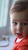 Lovely Caucasian kid standing at the window-sill. Baby makes a wry face moving forward to the camera. Close up. Blurred backdrop. Vertical video