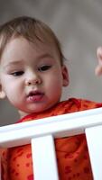Adorable baby boy standing in his cot looks at camera from top. Lovely kid notices something aside, smiles adorably and hurries there. Low angle view. Vertical video