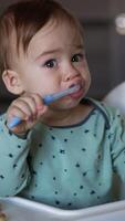 One-year old toddler sitting at the feeding table with spoon in mouth. Child turns to bagels left in front of him. Blurred background. Vertical video