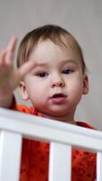 Cute one-year-old child standing in the crib and holding a pacifier in his hand. Baby looks straight into camera showing gestures with his hand. Vertical video