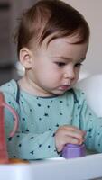 Cute adorable baby boy sitting in the feeding chair. Caucasian child with smeared face playing with a toy. Blurred backdrop. Vertical video