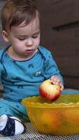 Lovely toddler sits on the floor with a yellow bowl in front of him. Cute kid takes an apple from a dish, looks at it carefully and turns it in his hands. Vertical video