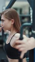 A beautiful girl with her strong arms performs an exercise on a simulator to strengthen the pectoral muscles in a fitness club. Close-up. Blurred Background Vertical video