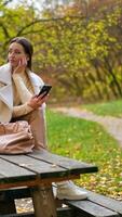 Thoughtful brunette lady sits on the table in the autumn park with phone in her hand. Woman answers the call and starts conversation. Blurred backdrop. Vertical video
