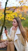 Attractive Caucasian woman using her phone sitting in the nature. Lady in warm clothes spending time in the autumn park and using gadgets and internet. Vertical video