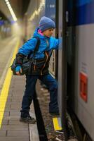 A schoolboy in a blue jacket and hat with a backpack enters a train carriage photo