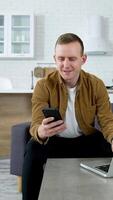 Smiling freelancer with a phone on kitchen background. Young man student using a laptop and mobile phone studying online. Guy working from home enjoying distant job. Vertical video