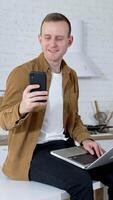 Portrait of a student with a laptop and a phone. Smiling young man is sitting on a kitchen table and using a laptop and mobile phone at home. Quarantine concept. Vertical video