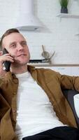 Young man talking the phone in the kitchen. Guy is sitting on sofa with white mug and speaking on a mobile phone at home. Vertical video