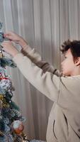 Beautiful high Christmas tree decoration in the house. Teenage boy looks at the toy snowflake with a smile and then puts it on the tree. Vertical video