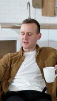 Handsome man using a phone at home. Guy rests on sofa with a cup of coffee and doing photos on a smart phone in the kitchen. Vertical video