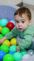 Lovely kid plays at the dry pool filled with colorful toys. Small child plays indoors. Top view. Vertical video