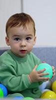 Sweet Caucasian kid sits inside the dry pool. Baby picks one ball, holds it and drops looking into camera. Vertical video