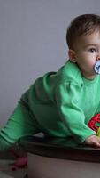 Caucasian toddler with pacifier in mouth sits on the stairs. Kid takes a toy, drops a pacifier and pulls the toy to the mouth. Vertical video