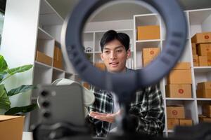 Young entrepreneur presenting product online using smartphone and ring light in modern workspace filled with boxes. photo