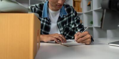 Young entrepreneur taking notes for online sales strategy, writing in notebook at modern workspace with product boxes. photo