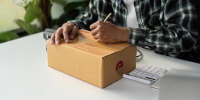 Person writing on cardboard box while preparing for online order shipment with calculator nearby. photo