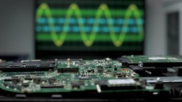 A circuit board is placed in the foreground while an oscilloscope in the background displays fluctuating electronic signals. The setting is a modern laboratory. video