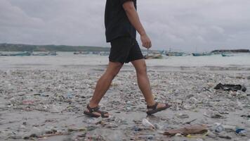 Man on Black Shirt Walking Ocean Plastic Waste Pollution, Dirty Polluted Trash Beach in Kedonganan Bali Indonesia Environmental Crisis Side Feet video