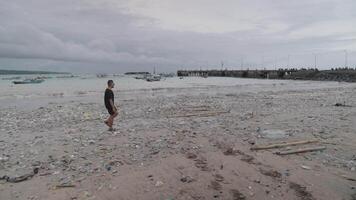 homem em Preto camisa caminhando oceano plástico desperdício poluição, sujo poluído Lixo de praia dentro kedonganan bali Indonésia de Meio Ambiente crise longe lado video