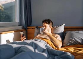 A young man enjoying a lazy Sunday morning, relaxing in bed with a cup of coffee and his phone photo