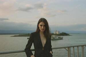 A young Caucasian woman , brown hair, wearing a black blazer with a deep neckline, standing by a railing. The background features a body of water with a boat photo