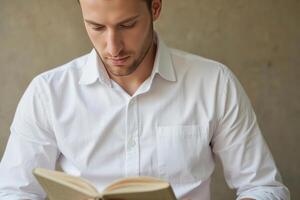 A caucasian man, age 28, with short brown hair, wearing a white shirt, reading a book in front of a gray wall photo