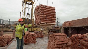 Construction worker guiding crane lifting bricks, A construction worker in a safety jacket and hard hat guiding a crane lifting a pallet of bricks. video