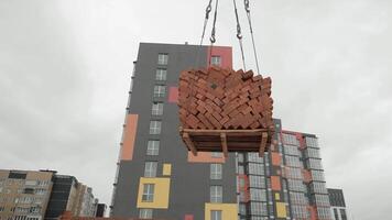 Crane lifting a pallet of bricks at a construction site, A pallet of red bricks being lifted by a crane near a modern building on a construction site. video