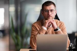 Smart looking focused businessman working on a laptop and thinking. Millennial managing family budget, handling financial paperwork, and tracking expenditures on a laptop at a modern open space office photo