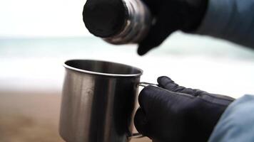 Unrecognizable woman pours water into a mug from a thermos near the sea, Slow Motion, closeup on a cup and thermos. The woman is wearing black gloves and a blue jacket. Cinematic frame. video