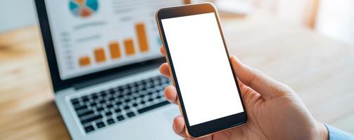 Marketing analytics performance ROI concept. Hand holding smartphone with a blank screen next to a laptop displaying charts. photo
