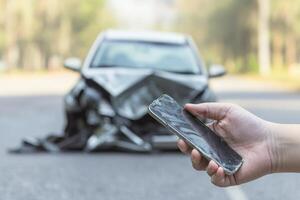 Vehicle with insurance and legal concept. Damaged phone held in front of a wrecked car on a road. photo