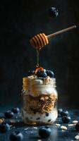 Layers of yogurt, granola, and blueberries in a glass jar with honey drizzling from a wooden stick photo