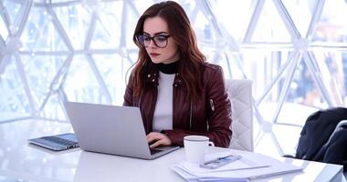 Woman working on laptop in modern office. Focused businesswoman seated at desk with documents and coffee, using a laptop. photo