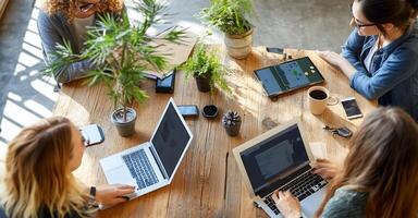 Women collaborating around a wooden table, surrounded by plants and technology, working on laptops and digital tablets. photo