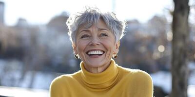 Portrait of a happy senior woman with short gray hair wearing a yellow turtleneck sweater, laughing and smiling at the camera. The background is blurred, with a sunny day and a bright white sky. photo