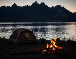 A tent is set up near a campfire on the shore of a lake photo