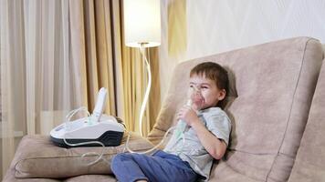 Child using nebulizer therapy. A young boy sits comfortably on a sofa, using a nebulizer to support his breathing while relaxing indoors. video