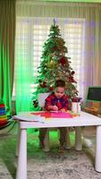 Child creating art by a decorated tree. A young child happily colors on bright paper at a table near a festive, decorated Christmas tree in a cozy room. video
