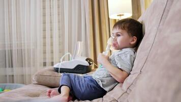 Child uses nebulizer at night. A young boy sits on a couch using a nebulizer while watching TV in a cozy living room setting during the evening. video