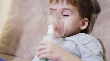 Child using nebulizer for breathing. A boy sits on a couch, using a nebulizer to administer medication for respiratory issues at home. video
