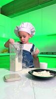 jovem chefe de cozinha preparando dentro uma brilhante cozinha. uma jovem criança dentro uma chefe de cozinha chapéu derrama farinha a partir de uma recipiente enquanto cozinhando dentro uma brilhante verde cozinha, criando uma bagunça. video