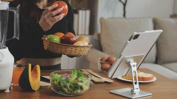 A woman holding a basket of fruit and vegetables while sitting at a table with a blender and other video