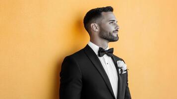 Groom in tuxedo poses against yellow wall photo