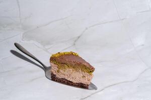 Slice of Dubai chocolate cheesecake with pistachio paste and kataifi covered in chocolate ganache on cake spatula. Marble background with copy space. Indulgence, gourmet desserts, culinary photo