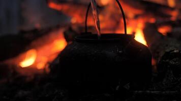 Water is being poured into a black kettle resting on a campfire. The warm glow from the flames illuminates the surrounding area, creating a cozy atmosphere in nature. video