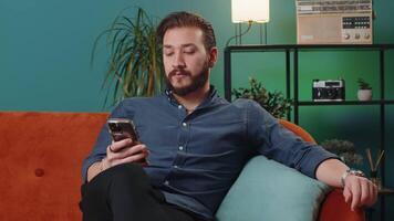 Lebanese arabian man sitting on sofa, using smartphone share messages on social media application video