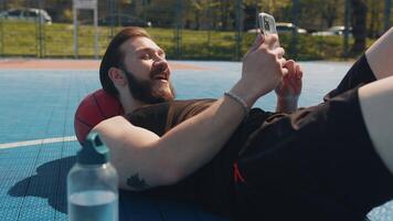 Tired man in sportswear after playing basketball game lying down on ball relaxing using smartphone video